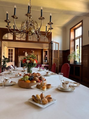 Ein Esszimmer mit einer Kronleuchter, Holzvertäfelung, einem großen Esstisch und Blick auf einen Garten durch ein Fenster.