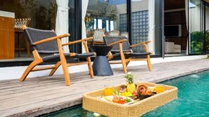 Ein Poolbereich mit zwei Korbstühlen, einem Korbstuhl-Tisch mit Obst- und Getränkeplatte, und einem modernen Gebäude im Hintergrund.