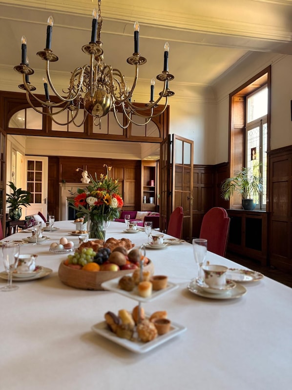 Une salle à manger agrémentée d’un lustre, de boiseries, d’une grande table dressée pour le repas, et offrant une vue sur un jardin par la fenêtre.
