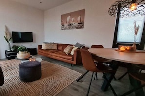 A modern living room with a sofa, a dining table, and a TV mounted on the wall. - Holiday home in Olburgen with swimming pool (Olburgen)