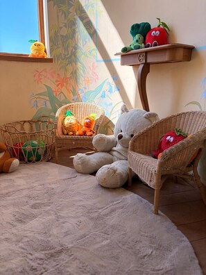 Een gezellige kamer met een groot teddybeer, rieten meubels en een wand met bloemmotieven.
