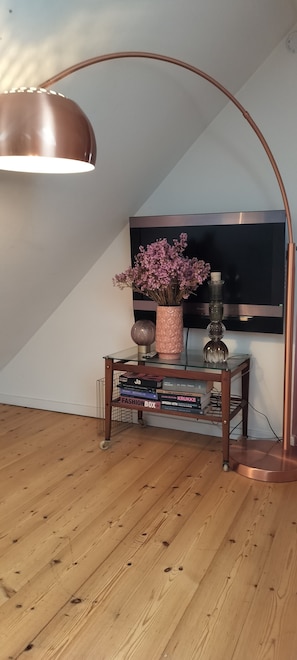 Et værelse med trægulv, et glastilbord med bøger, en vase med blomster, en lampe og et fjernsyn monteret på væggen.