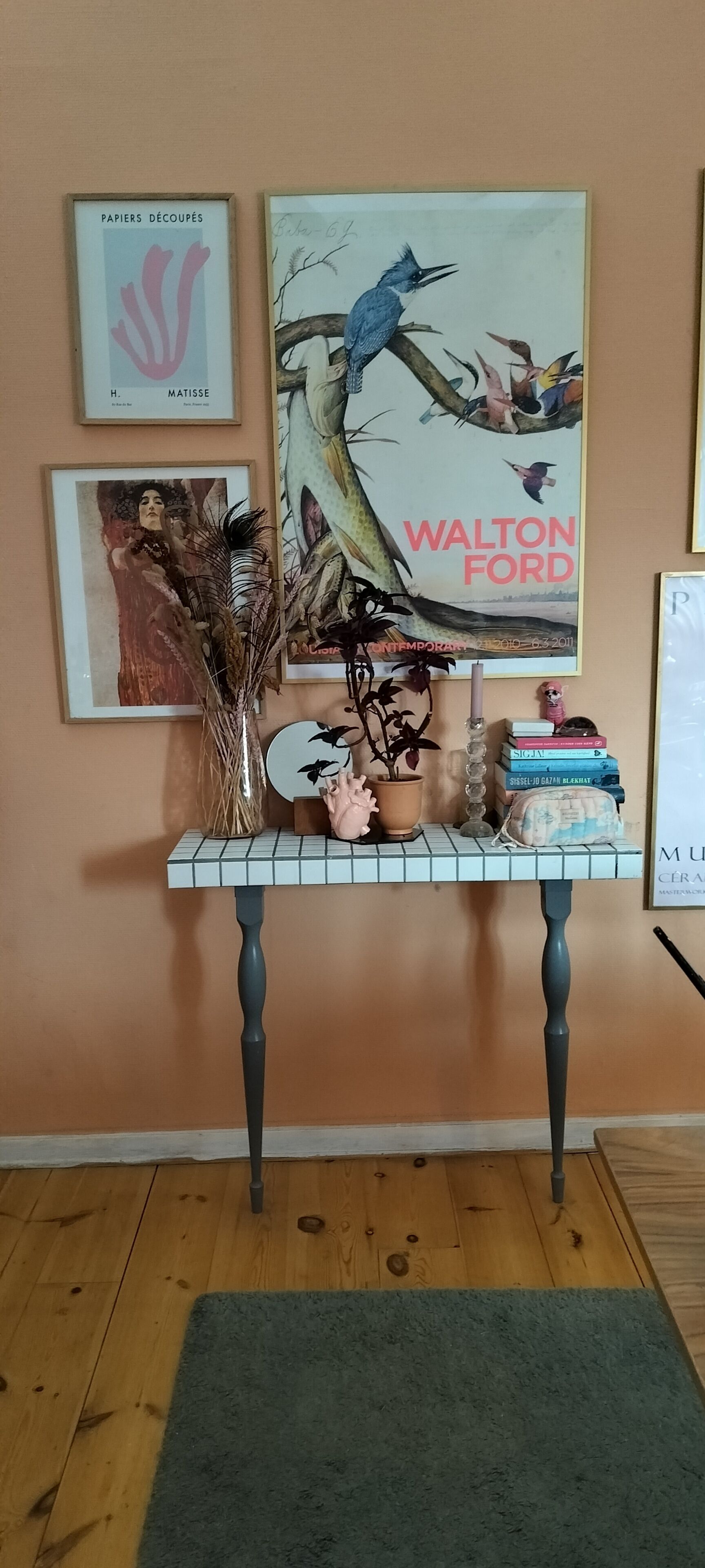 Een kamer met een houten vloer, een grijze vloerkleed, een tafel met een vaas, boeken en lijstwerken aan de muur.