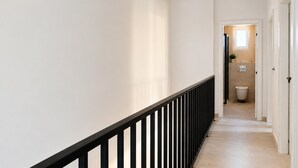 Un couloir étroit avec une rambarde noire, qui mène à une salle de bain équipée de toilettes et d’une petite fenêtre.