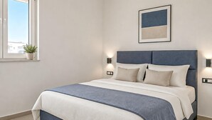 Une chambre à coucher bien rangée, avec un lit, des lampes de chevet, un tableau encadré au mur et une fenêtre agrémentée d’une plante.