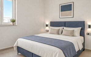 A neatly arranged bedroom with a bed, bedside lamps, a framed picture on the wall, and a window with a plant. - Nice home in Privlaka (Privlaka)