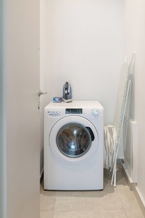 A compact, white front-loading washing machine in a small laundry room with a door, a wall-mounted drying rack, and a tiled floor. - Oddity Seafront Penthouse Apartments (Thessaloniki)