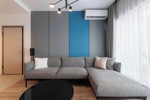 Un salon moderne avec un canapé d’angle, une table basse ronde et une grande fenêtre agrémentée de rideaux.