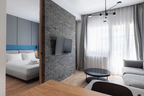 A modern living room with a grey brick accent wall, a flat-screen TV, a grey sofa, a round coffee table, and a wooden dining table. - Oddity Seafront Penthouse Apartments (Thessaloniki)