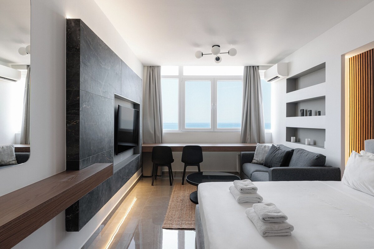 A modern hotel room with a large bed, a TV mounted on a dark wall, a desk with two chairs, and a view of the sea through large windows.