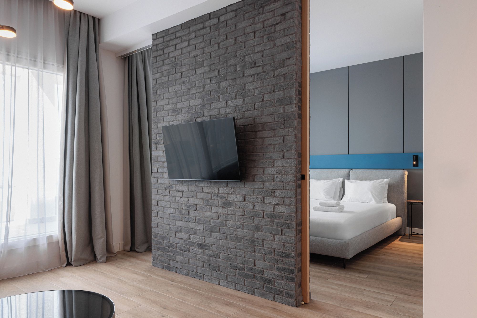 Une chambre d’hôtel moderne avec un mur d’accent en briques, un téléviseur à écran plat fixé sur ce mur, un lit avec une literie blanche et une touche de bleu sur la tête de lit.