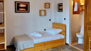 A single bed with a wooden headboard, a white pillow, and a neatly folded towel. There are two framed pictures on the wall, a wooden shelf with a red item, and a white toilet visible. - Oddity Seafront Penthouse Apartments (Thessaloniki)