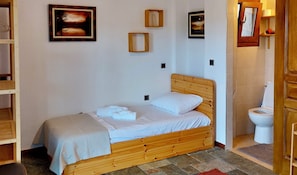 Ein Einzelbett mit Holz Kopfteil, einem weißen Kissen und einem ordentlich gefalteten Handtuch. An der Wand hängen zwei gerahmte Bilder, und ein Holzregal mit einem roten Gegenstand ist zu sehen. Außerdem ist eine weiße Toilette sichtbar.