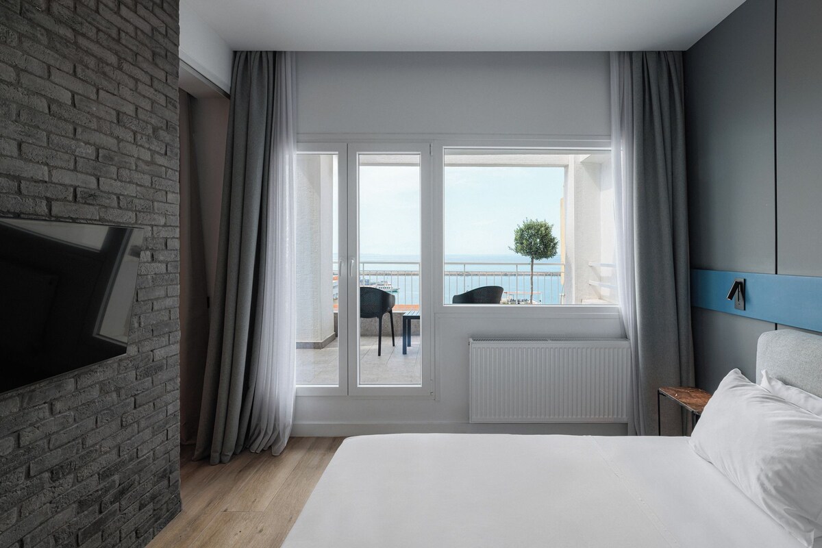 A hotel room with a large bed, a view of the sea, a balcony with a table and chairs, and a brick wall.