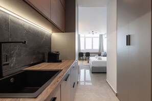 Une cuisine moderne équipée d’une cuisinière intégrée, d’une machine à café et d’un évier. Un couloir mène à une chambre à coucher qui comprend un lit et une fenêtre.