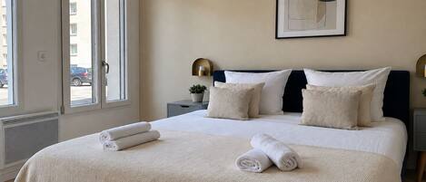 A bedroom with a large bed, a nightstand, a wall-mounted lamp, and a window with a view of the outside.