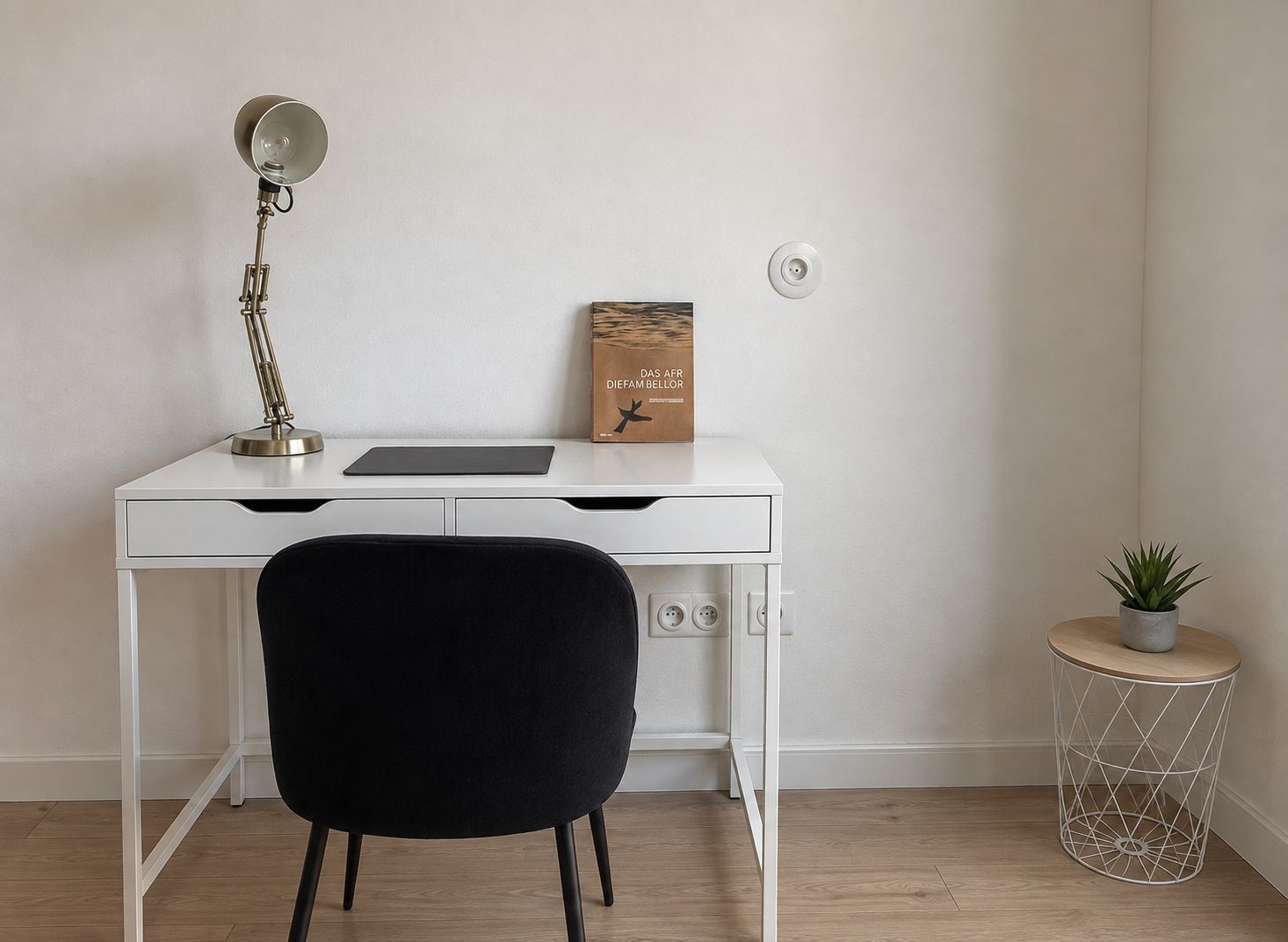 Un bureau blanc avec une lampe, une boîte étiquetée « DIEFAMBELLOR », une chaise noire et une petite table ronde avec une plante.