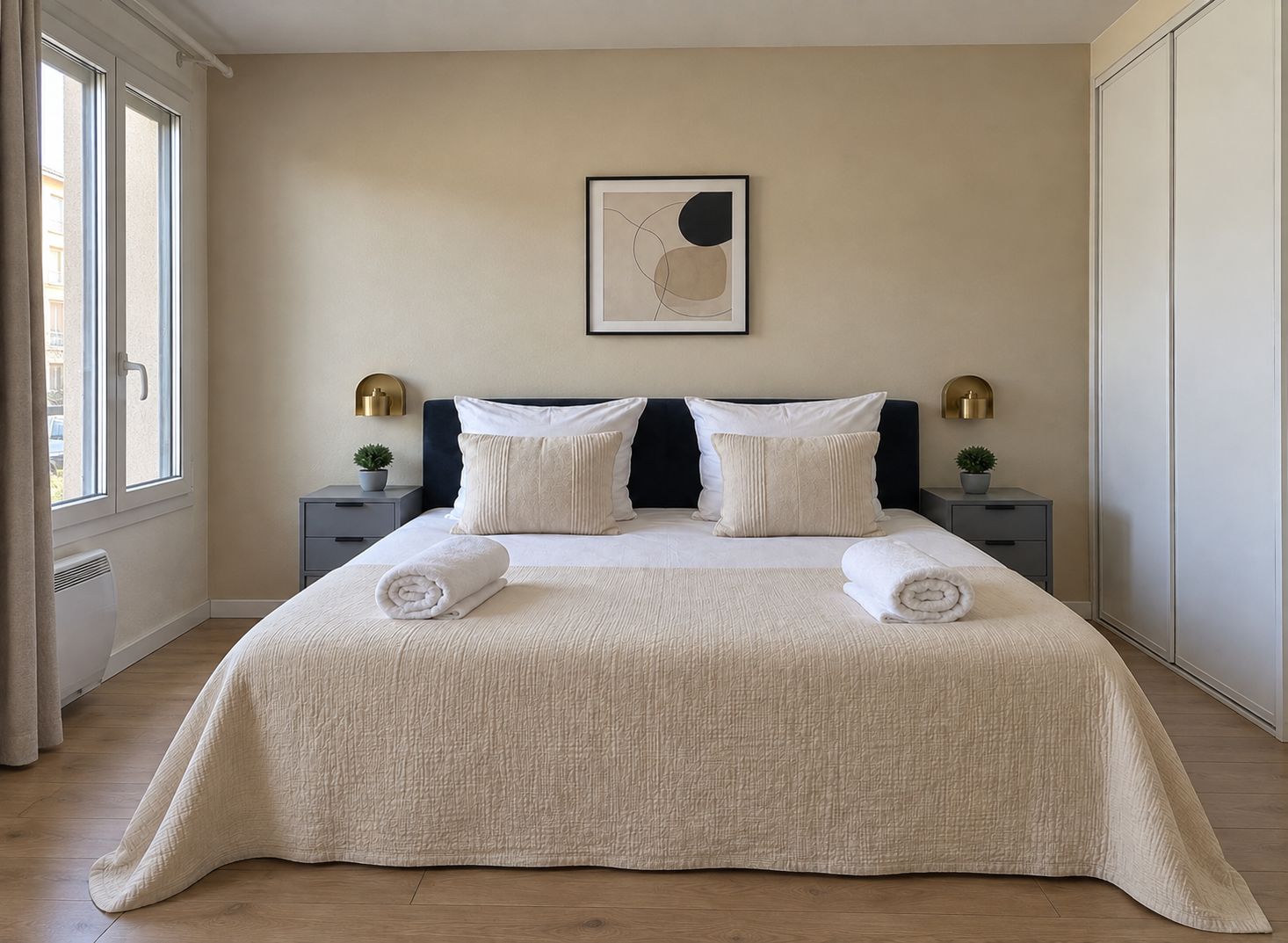 Una cama bien tendida con una colcha texturizada, dos almohadas y dos toallas enrolladas. Hay una obra de arte colgada en la pared sobre la cama, y dos mesitas de noche con lámparas.
