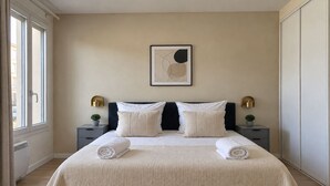 A neatly made bed with a textured bedspread and two rolled towels, a wall-mounted abstract artwork, bedside lamps, and a potted plant on a nightstand. - Magnifique 2 pieces a Issy lesMoulineaux (Vanves)