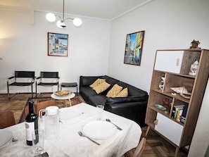 Een eetgedeelte met een tafel gedekt voor twee personen, een zwarte leren bank, twee zwarte stoelen, een boekenkast en twee schilderijen aan de muur.