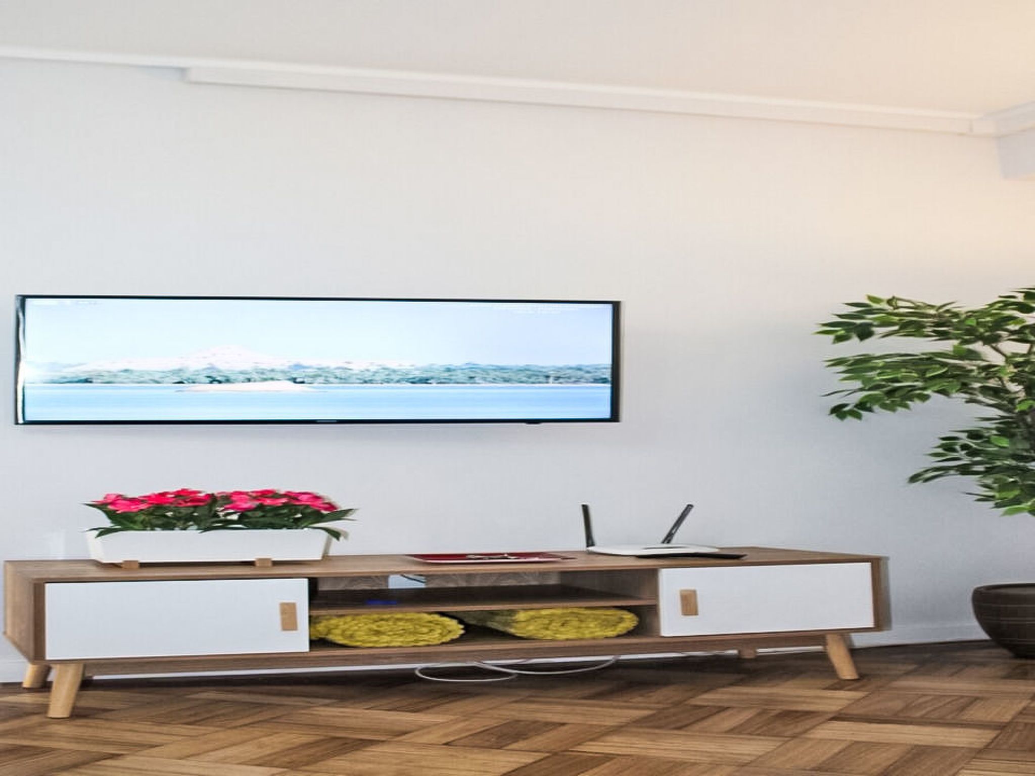 En moderne stue med et fladskærms-tv monteret på væggen, et træsofa bord med skuffer, en potteplante og en stor grøn plante.