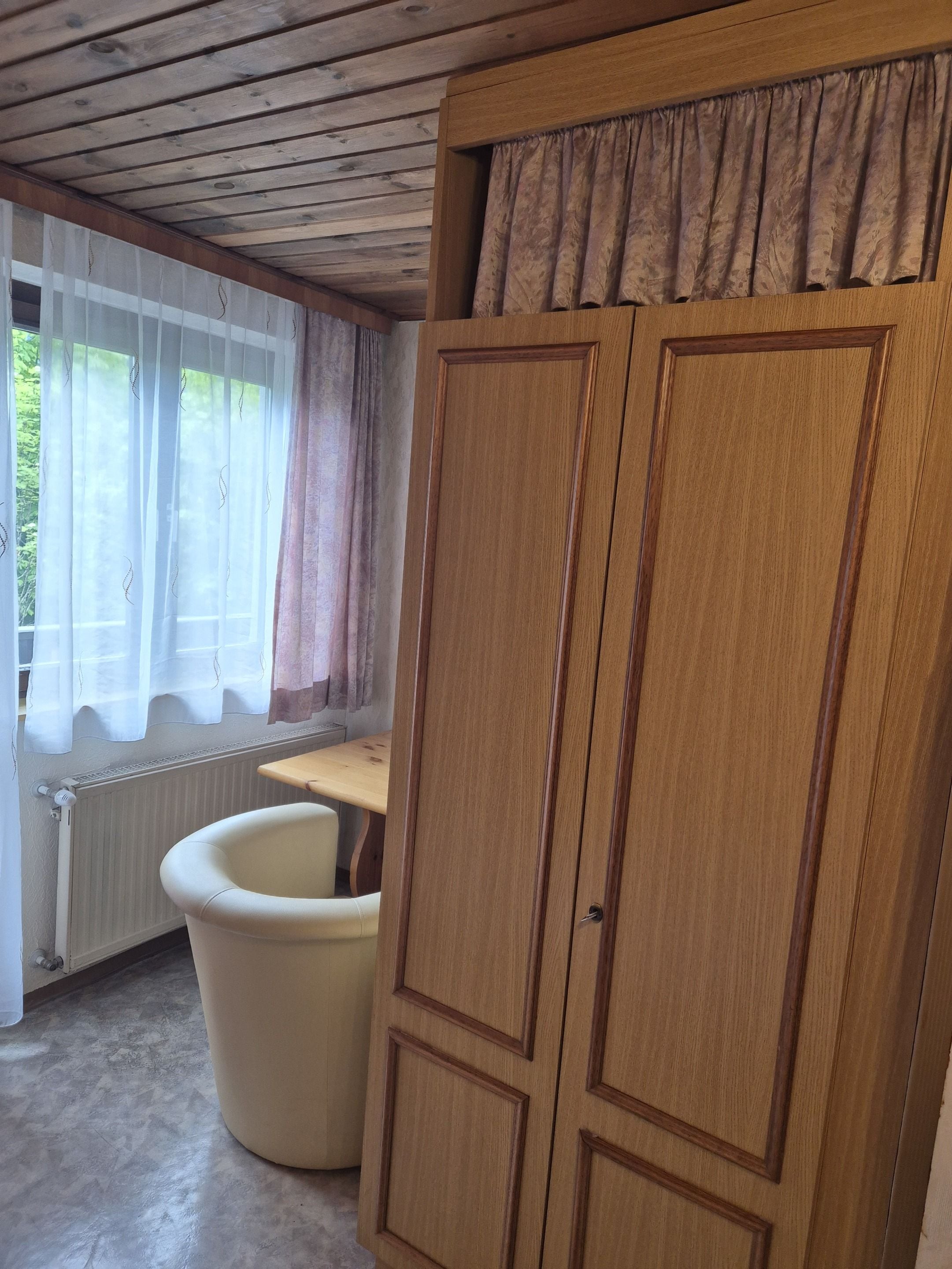 Una habitación con un armario de madera, una silla blanca, una mesa y un ventanal con cortinas.