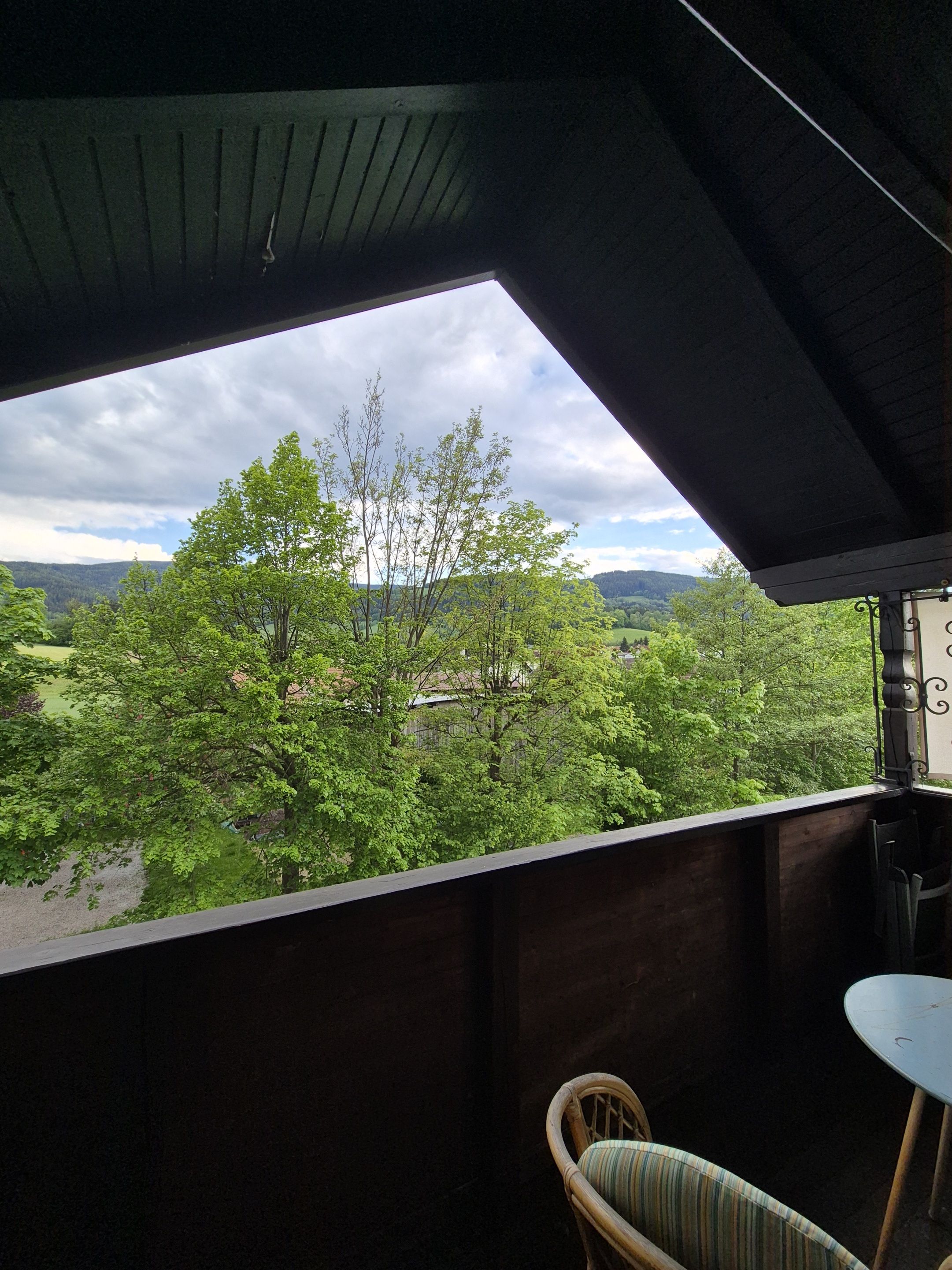 A balcony with a view of a green landscape and trees, a wooden ceiling, and a small table with a chair.