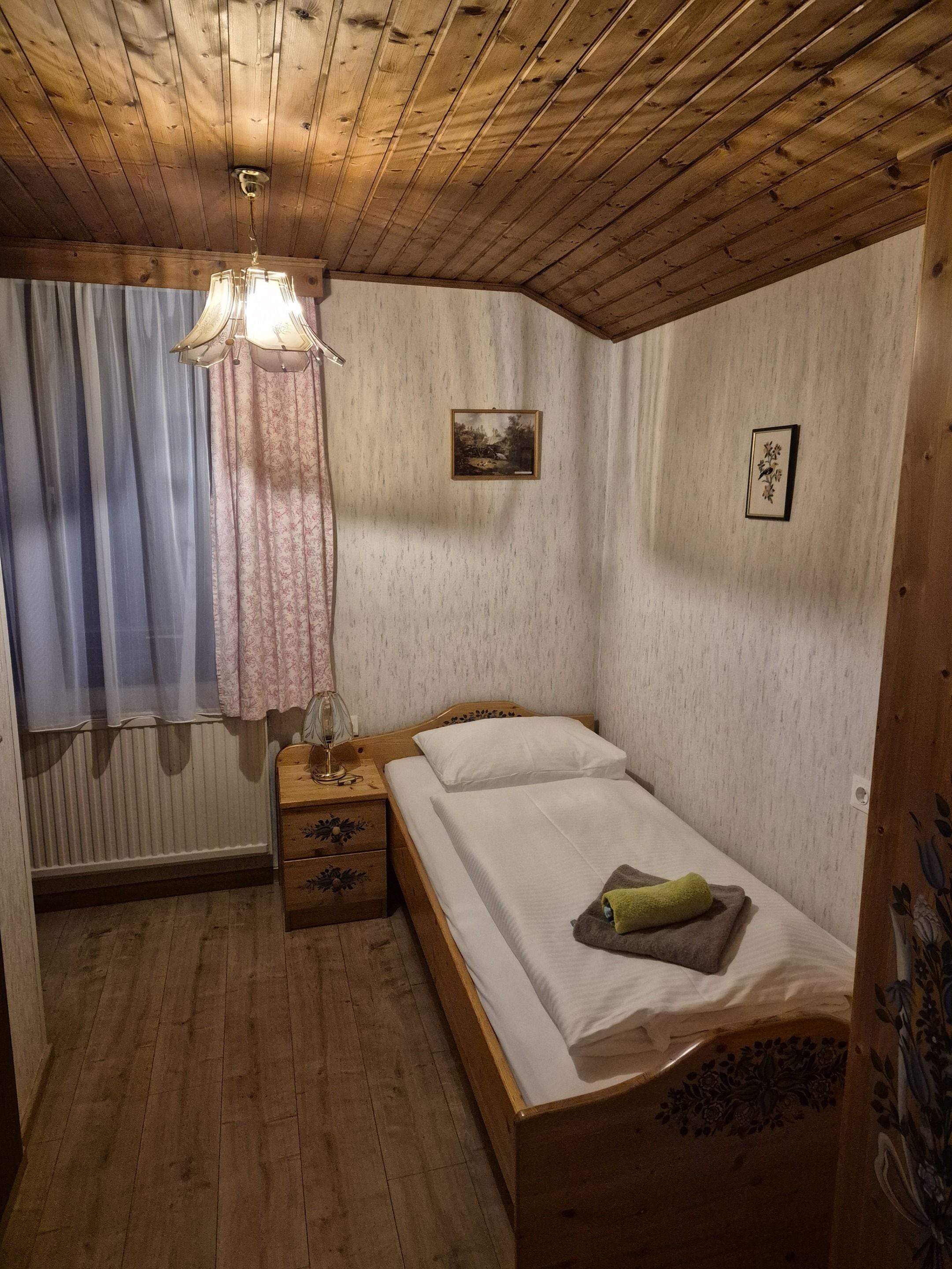 A small, cozy bedroom with a wooden ceiling, a single bed, a nightstand, a window with curtains, and a radiator.