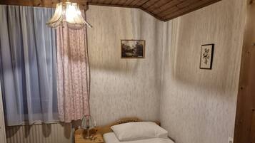 A small, cozy bedroom with a wooden ceiling, a single bed, a nightstand, a window with curtains, and a radiator.