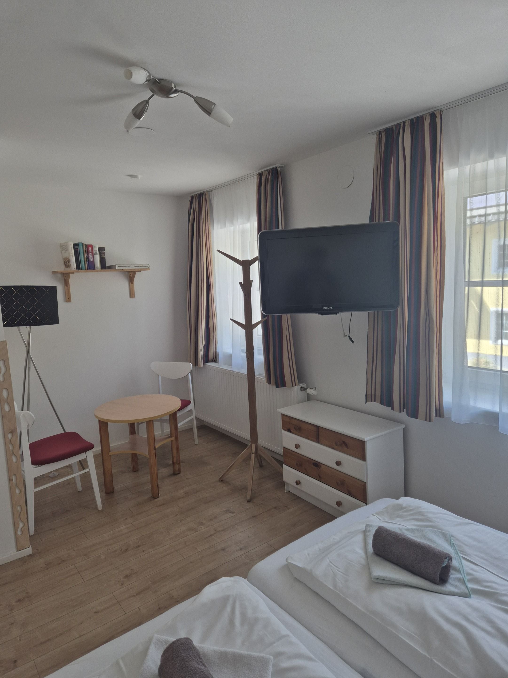 A hotel room with a bed, a desk with a chair, a television, a radiator, and a window with curtains.