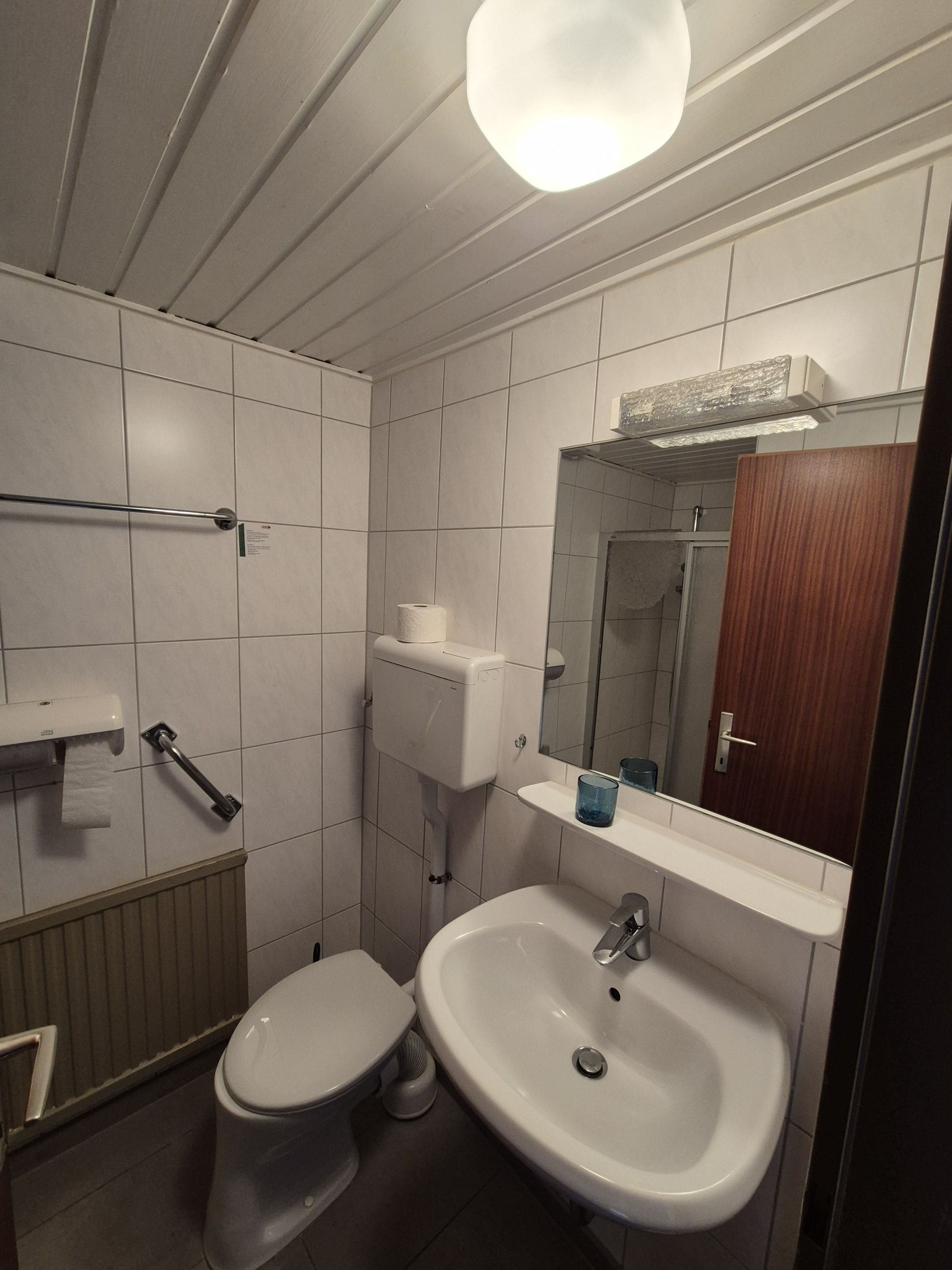 Baño con inodoro, lavamanos, espejo y dispensador de toallas.