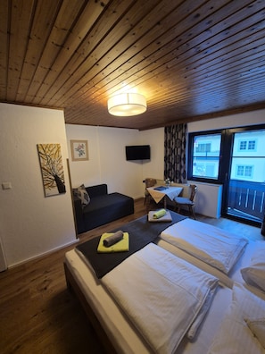Una habitación con techo de madera, una cama con ropa blanca, un sofá pequeño, una mesa con una silla y una ventana con vistas a edificios.