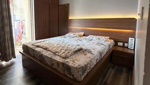 A bedroom with a bed, a ceiling fan, a nightstand, a wardrobe, and a window with curtains. - The Nest Noida (Noida)