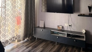 A modern living room with a flat-screen TV, dark wood flooring, a ceiling fan, and patterned curtains. - The Nest Noida (Noida)