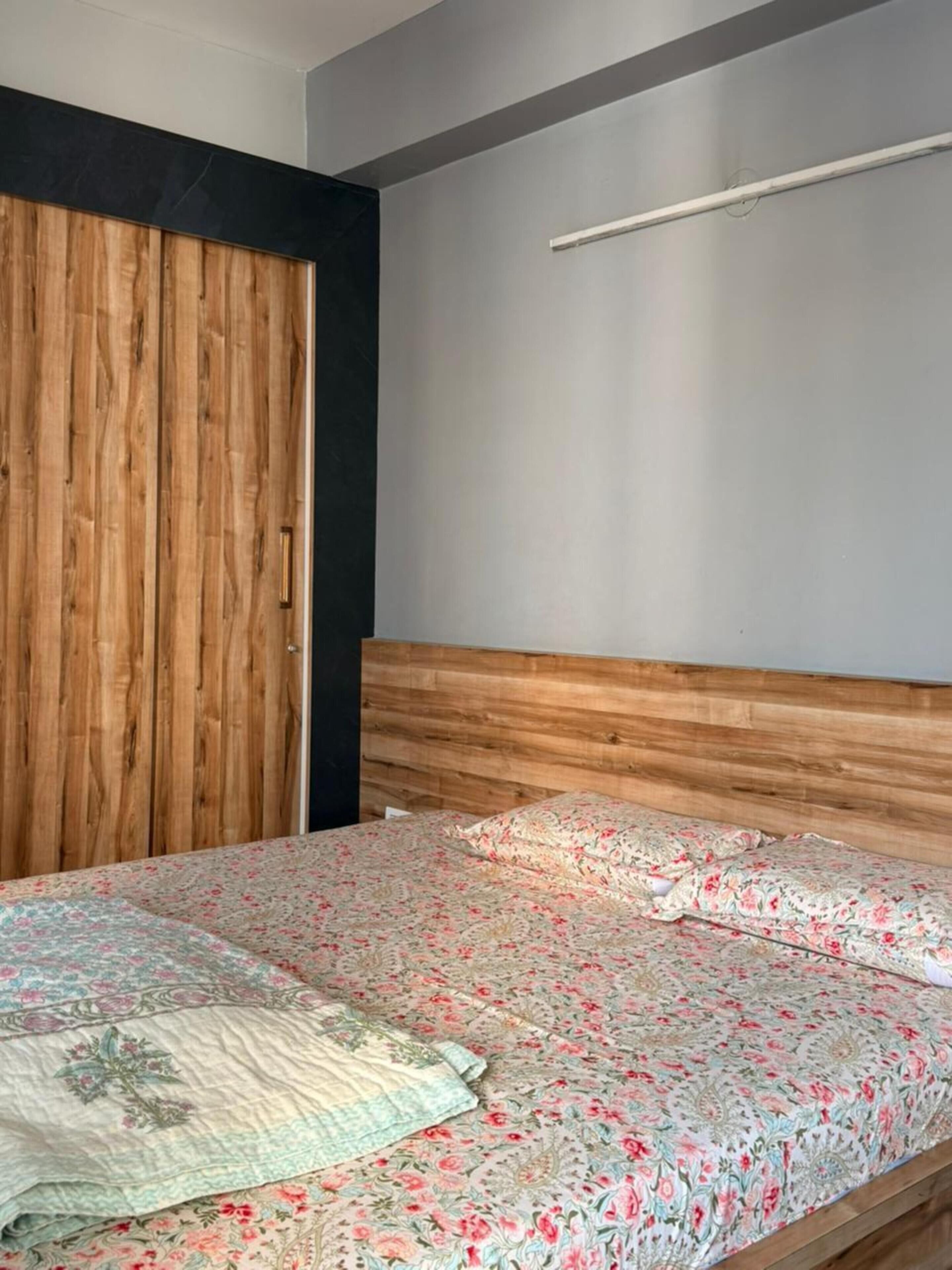 A bedroom with a wooden wardrobe, a bed with floral bedding, and a plain wall with a light fixture.