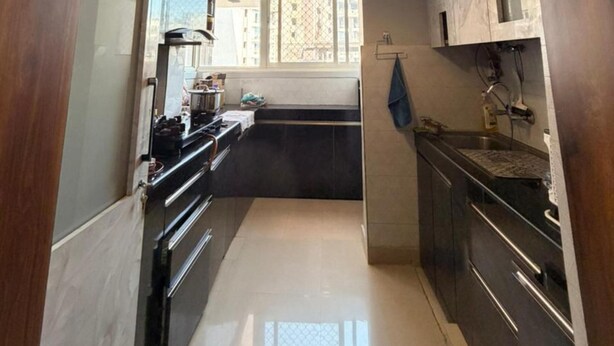 A narrow kitchen with dark cabinets, a central island, and a hanging pendant light. - The Nest Noida (Noida)