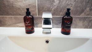 En handfat i badrummet med två flaskor av Odyssey Black Pepper & Sandalwood-schampo och duschgel, en kromad kran och en vägg täckt med marmorplattor.