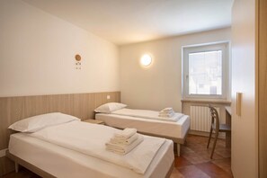 A hotel room with two beds, a desk, a chair, a radiator, and a window with blinds. - Casa Gloria - in the center of San Zeno di Montagna (San Zeno di Montagna)