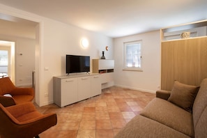 Ein Wohnzimmer mit Fernseher, einer Couch, einem Sessel, einem Schrank und einem Fenster.