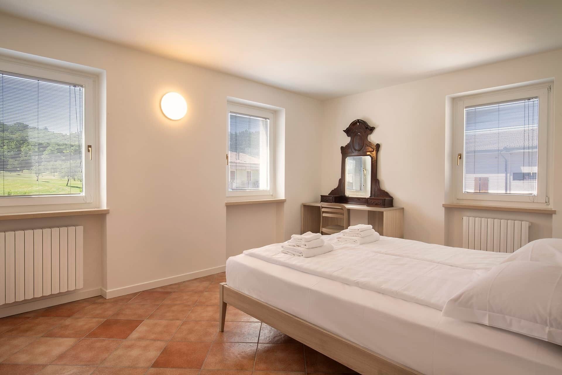 Ein Schlafzimmer mit einem Bett, einer Kommode, einem Spiegel und zwei Fenstern mit Jalousien.