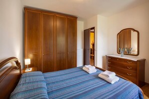 A bedroom with a large wooden wardrobe, a bedside table with a lamp, a dresser with a mirror, and a bed with blue-striped bedding and towels. - Casa Lisa - tranquility in San Zeno (San Zeno di Montagna)