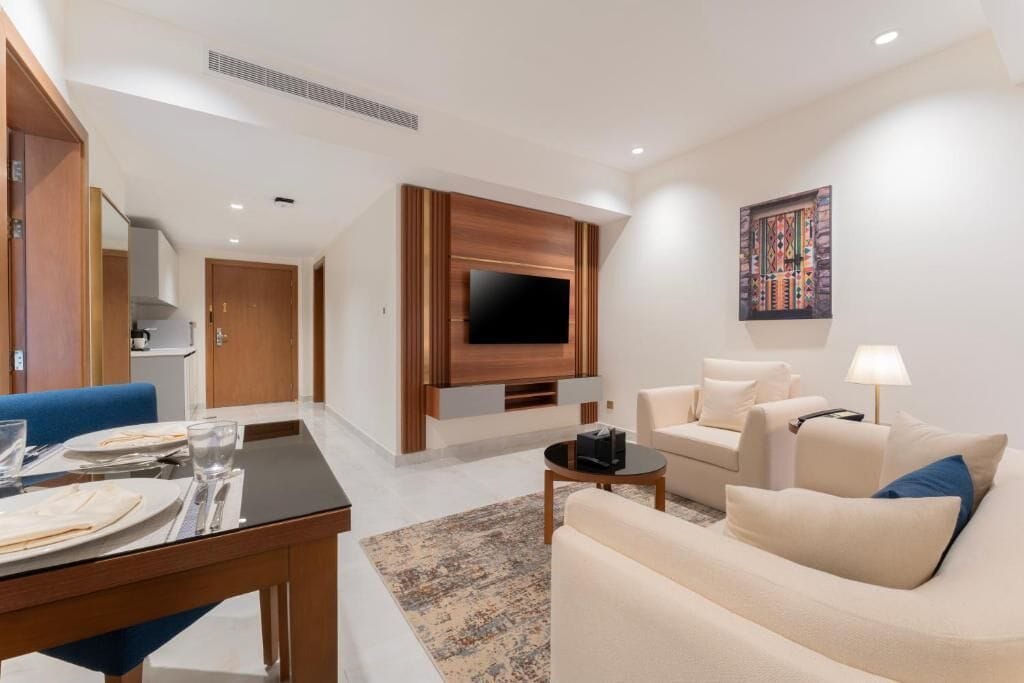 A modern living room with a flat-screen TV mounted on a wooden panel, a sofa, a coffee table, and a dining area with a table set for two.
