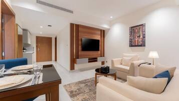 A modern living room with a flat-screen TV mounted on a wooden panel, a sofa, a coffee table, and a dining area with a table set for two.