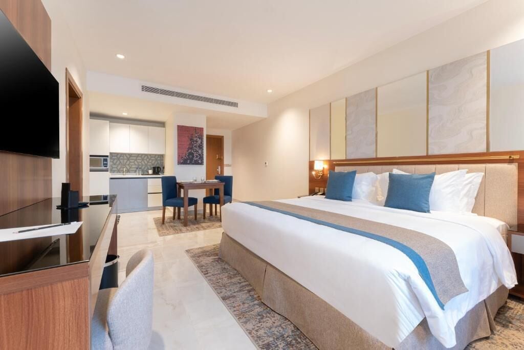 Quarto de hotel com uma cama grande, uma escrivaninha, televisão e uma cozinha compacta ao fundo.