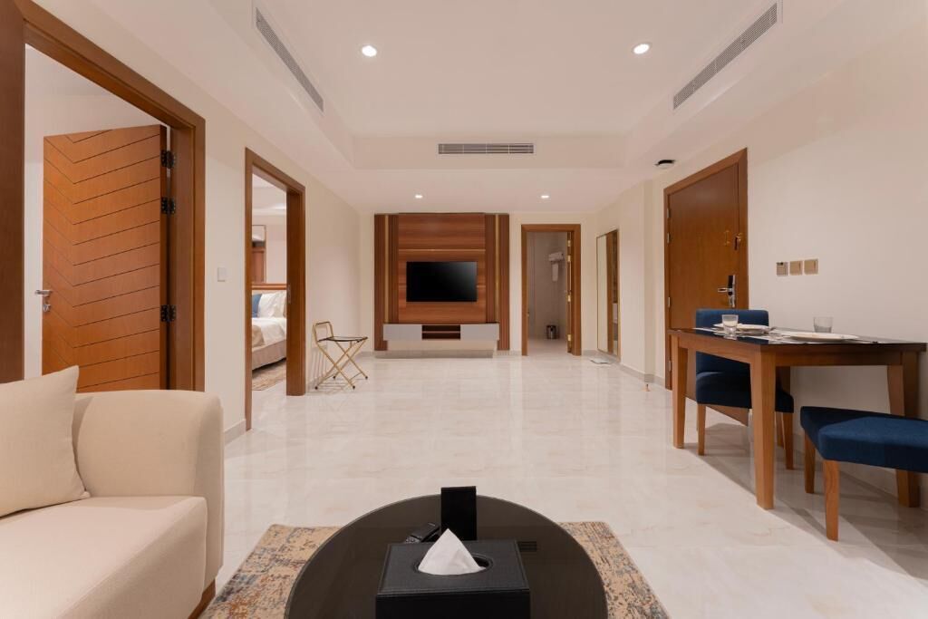 A hotel room with a sofa, a dining table with chairs, a TV mounted on the wall, and a small table with a tissue box.