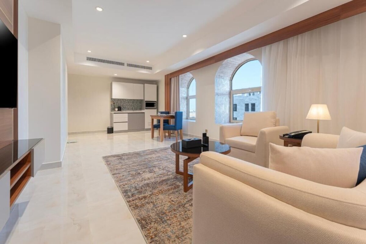 Een moderne woonkamer met een bank, fauteuils en een salontafel. Achtergrond is een keuken met een eethoek.