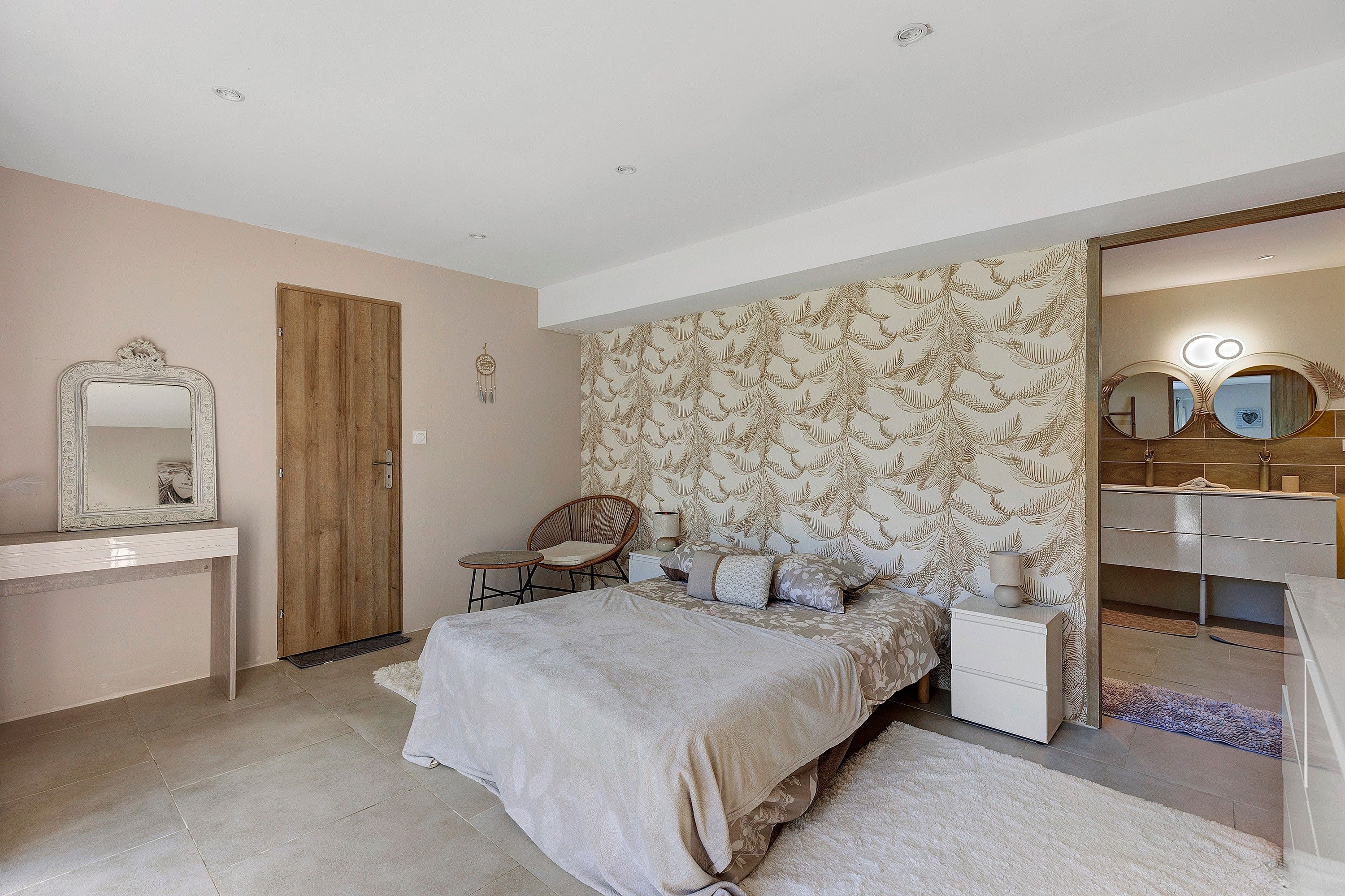 A bedroom with a bed, a chair, a nightstand, a mirror, a wardrobe, and a bathroom with a sink and mirror.
