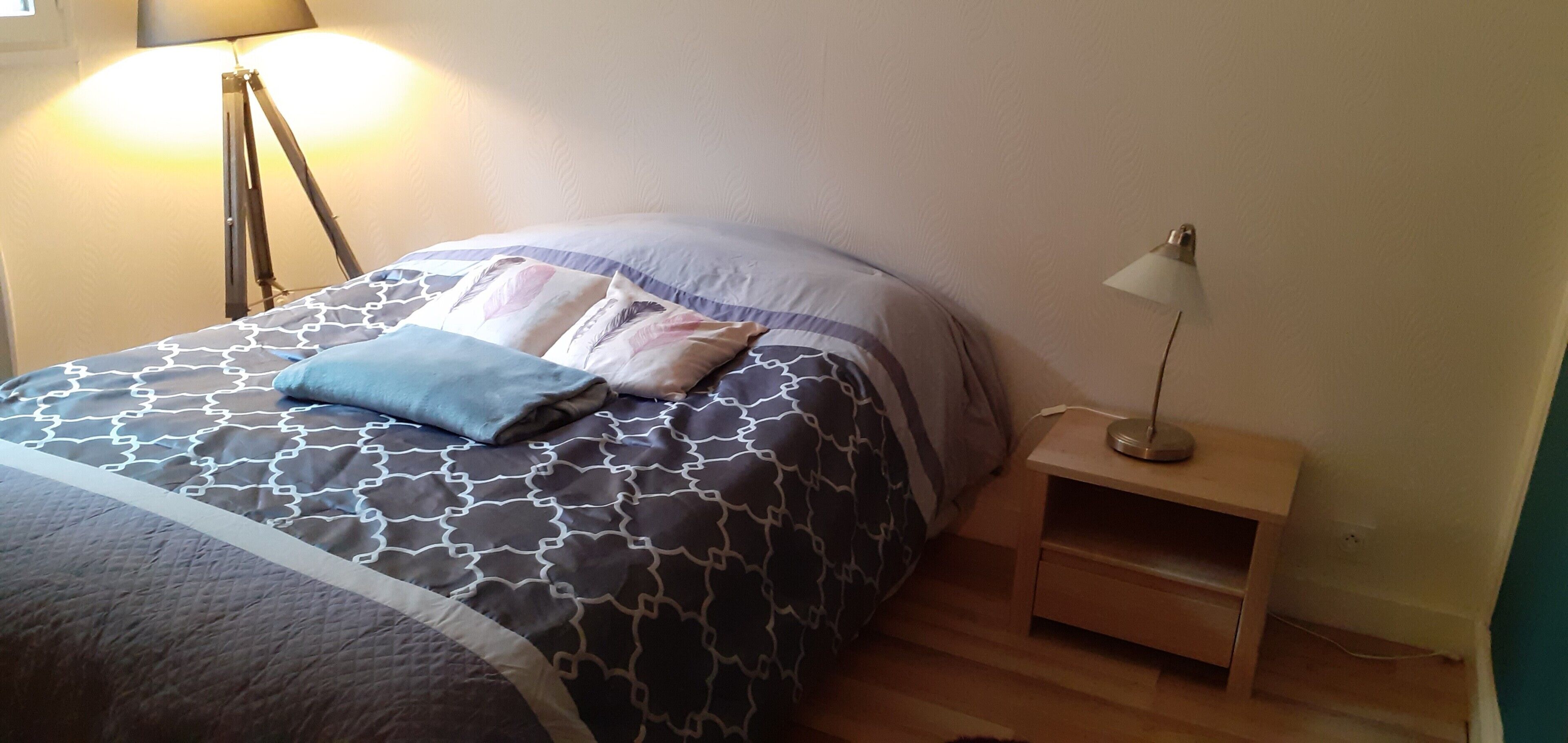 Una cama bien hecha con ropa de cama estampada, una mesita de noche con lámpara y una estantería en la pared.
