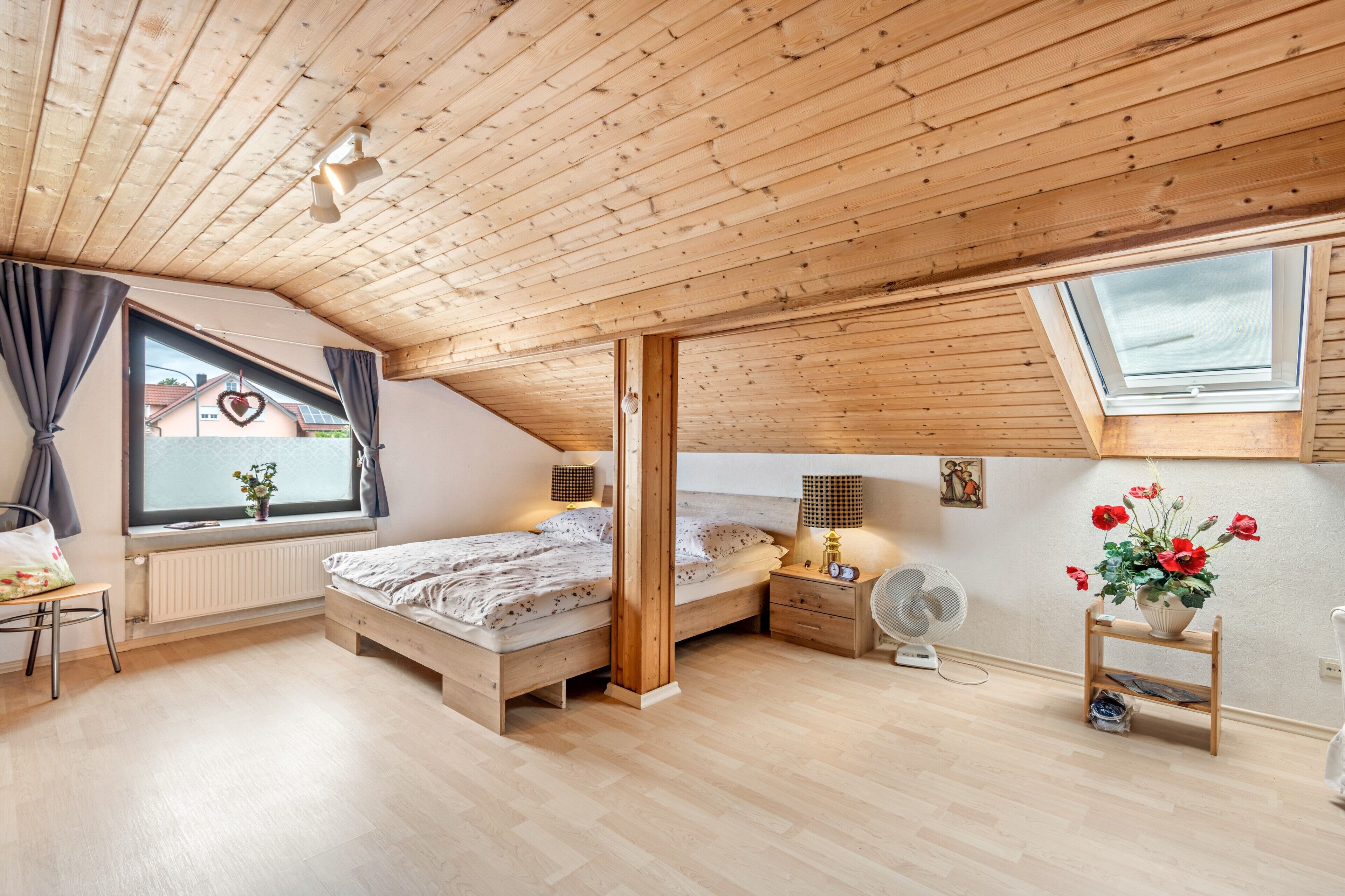 A bedroom with a sloped ceiling, wooden beams, a window with curtains, a bedside table with a lamp, a fan, and a small shelf with flowers.