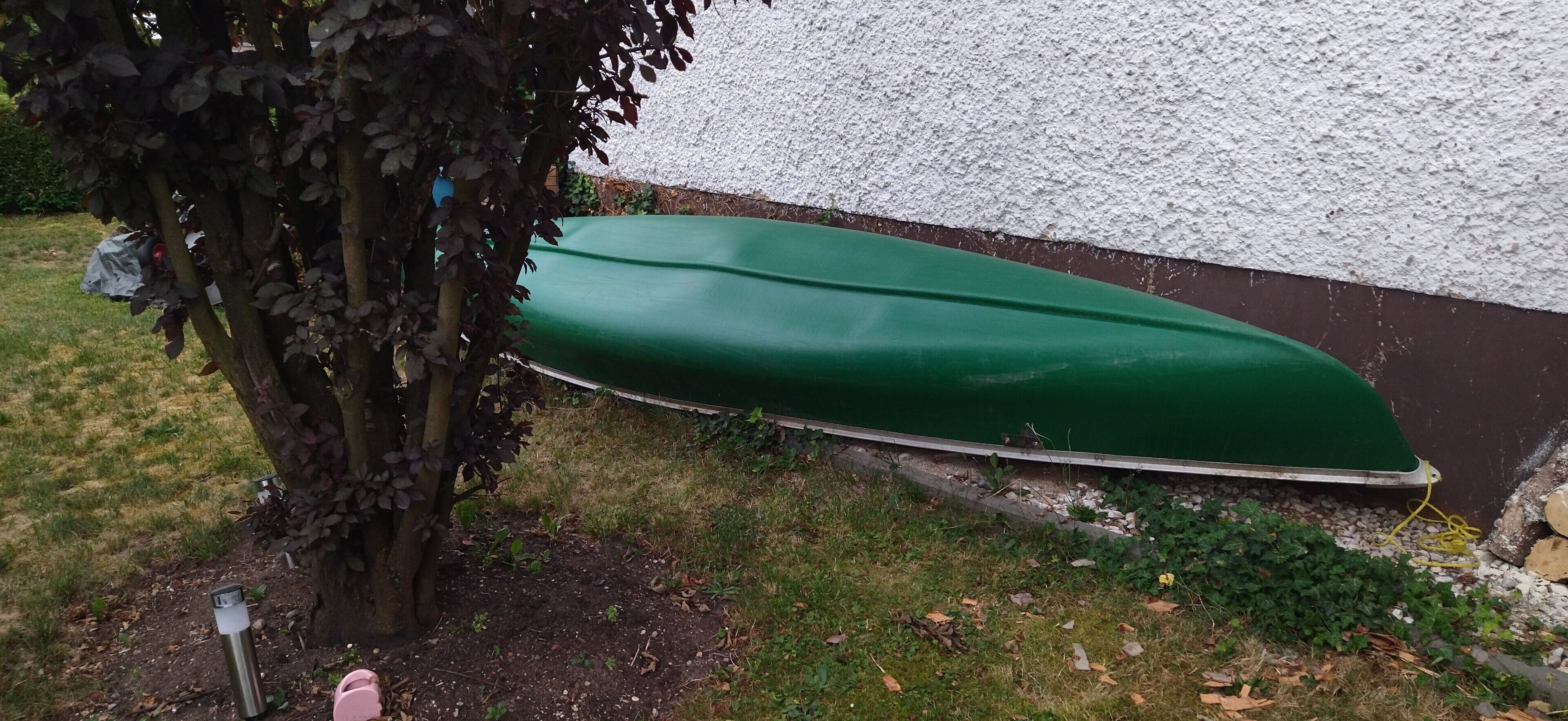 Un bateau vert, recouvert d’une bâche, partiellement dissimulé par un arbre, avec un mur blanc à texture visible en arrière-plan.